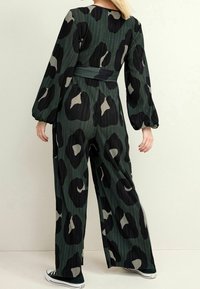 Combinaison à imprimé léopard vert avec texture plissée, manches longues bouffantes et taille ceinturée. Des accents noirs et beiges sont présents.