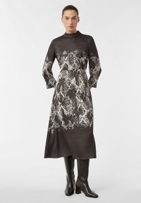 Langärmliges Kleid aus dunklem Stoff mit einem weißen Schlangenhautmuster, hohem Kragen und lockerer Passform, kombiniert mit knielangen schwarzen Stiefeln.
