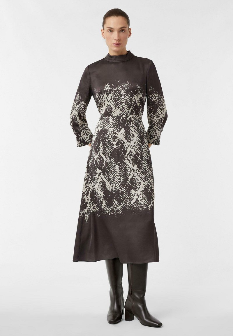 Langärmliges Kleid aus dunklem Stoff mit einem weißen Schlangenhautmuster, hohem Kragen und lockerer Passform, kombiniert mit knielangen schwarzen Stiefeln.