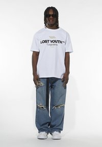 Biely oversized tričko s čiernym logom "LOST YOUTH", v kombinácii so zničenými modrými džínsami a bielymi teniskami. Doplnkové slnečné okuliare.