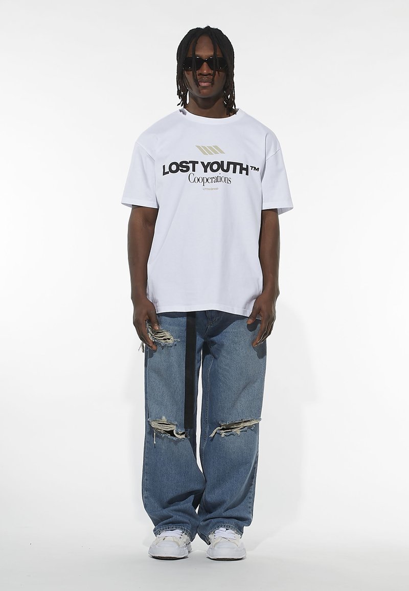 Biely oversized tričko s čiernym logom "LOST YOUTH", v kombinácii so zničenými modrými džínsami a bielymi teniskami. Doplnkové slnečné okuliare.