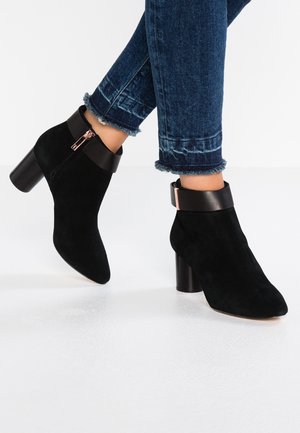 Person iført mørkeblå jeans med frynset kant og sorte ruskind ankelstøvler med blokhæl og sidelommer med lynlås.