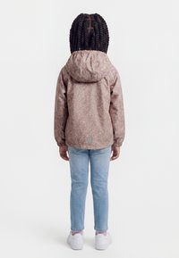 Copil cu păr lung împletit, purtând o jachetă cu glugă cu model, blugi albastru deschis și adidași albi, stând cu fața spre spate pe un fundal alb.