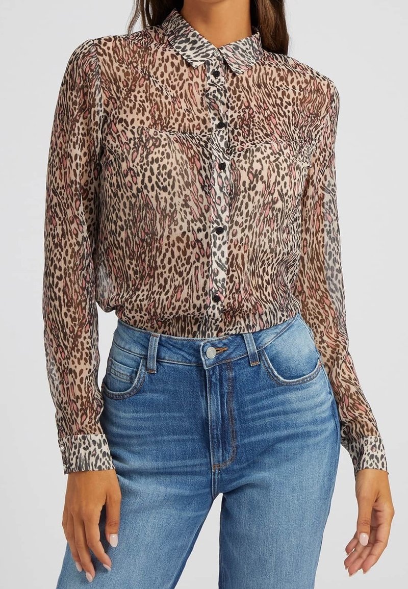 Femme portant un chemisier transparent à manches longues avec imprimé animal, rentré dans un jean bleu taille haute avec boutons et poches à l'avant.