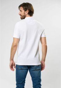 White short-sleeve polo shirt with a classic collar, smooth texture, and side vents. Worn with blue denim jeans, view from the back.