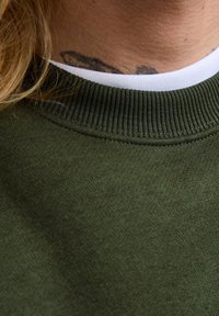 Närbild av en nacke med en mörk abstrakt tatuering ovanför den vita skjortkragen under en grön tröja, med långa blonda hårstrån synliga.