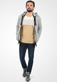 Grå zip-hoodie med sorte snore over en beige og grå stribet t-shirt. Mørkeblå jeans og grå sneakers fuldender outfittet.
