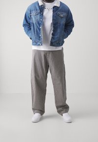 Man som bär en blå denimjacka med vit shearlingkrage, vit graphic t-shirt, randiga lösa byxor och vita sneakers, stående med händerna i fickorna.