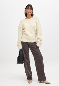 Krämfärgad oversized stickad tröja med ribbade muddar och nederkant, ihop med mörkbruna vida byxor. Modellen håller en svart handväska och har på sig spetiga beigefärgade skor.