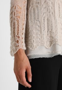 Ljust beige stickad tröja med öppen spetsmönster, texturerade ärmar och fransad särkant, lagrad över en vit genomskinlig blus.