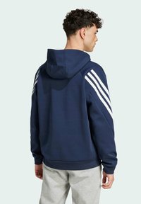 Námořnicky modrá mikina s kapucí a bílým designem tří pruhů na ramenou. Vyrobena z měkké tkaniny, má žebrované manžety a klokaní kapsu.
