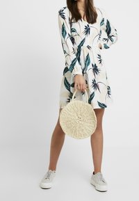 Blumenkleid in Creme mit grünen und dunkelblauen Blättern, tailliert, lange Ärmel; hält eine runde, geflochtene Strohtasche.