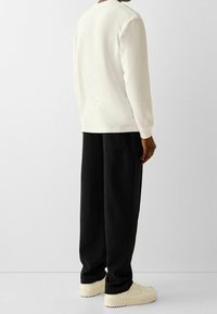 Person som bär en långärmad vit skjorta, lösa svarta byxor och off-white sneakers, som står mot en enkel ljus bakgrund och vänder ryggen åt.