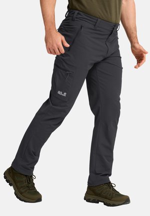 Man die donkergrijze outdoorbroek met ritszakken en olijfgroene wandelschoenen draagt, bij de taille afgesneden.
