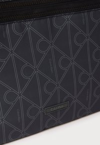 Ciemnoszara torba Calvin Klein z geometrycznym wzorem i czarnym zamkiem błyskawicznym, z małą prostokątną etykietą Calvin Klein.