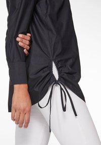 Schwarze Hemdbluse mit einem Kordelzugdetail in der Taille, langen Ärmeln und einem glatten, leicht strukturierten Stoff.