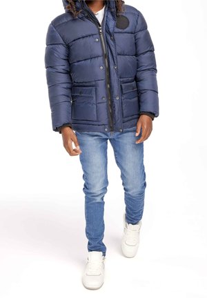 Personne marchant portant une doudoune bleu marine, un jean bleu clair et des baskets blanches sur un fond blanc.