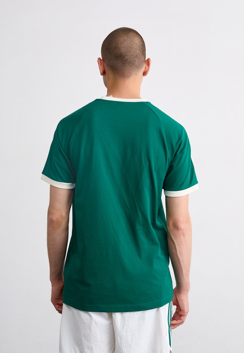 Camiseta de algodón verde con mangas raglán cortas, con un cuello redondo blanco y puños de las mangas. Textura suave, diseño sencillo.