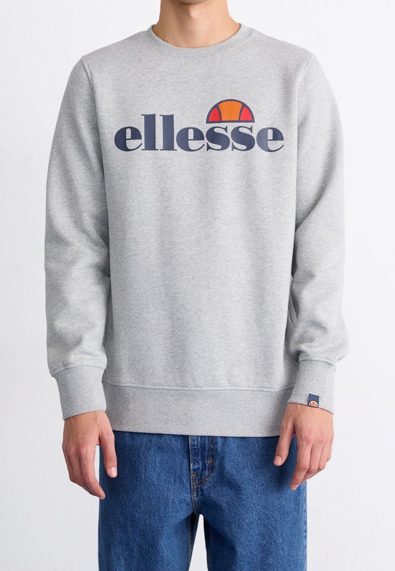 Homme portant un sweat-shirt gris clair avec le logo "ellesse" en bleu marine et orange, associé à un jean bleu, debout devant un fond blanc.