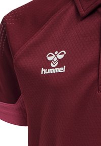 Mörkröd pikétröja tillverkad av strukturerad tyg, med en vit logotyp på vänster bröstområde och en krage med en något mörkare kant.