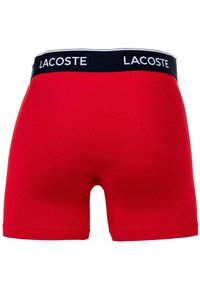 Czerwone bawełniane bokserki z czarną gumką w talii, na której znajduje się białe logo "LACOSTE". Gładka tekstura i dopasowany krój.
