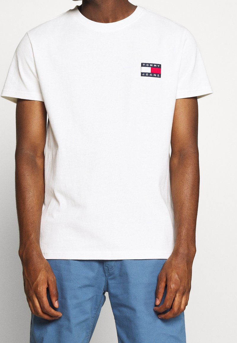 Bílý tričko s krátkým rukávem a kulatým výstřihem. Na levé straně hrudi se nachází barevná nášivka s logem "Tommy Jeans". Jednoduchá bavlněná textura.