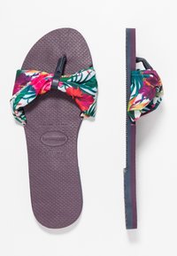 Flip-flop sandal med en lila gummisula, med en färgglad blommig överdel i tyg och en detalj i form av en rosett, samt en texturerad innersula för bra grepp.