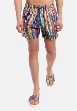 Homme portant un short à motifs colorés et des claquettes assorties, marchant vers l'avant, montré de la taille aux pieds sur un fond blanc uni.