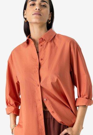 Chemise orange à manches longues avec une coupe décontractée, manches retroussées et une texture lisse. Présente un col classique et un léger éclat.