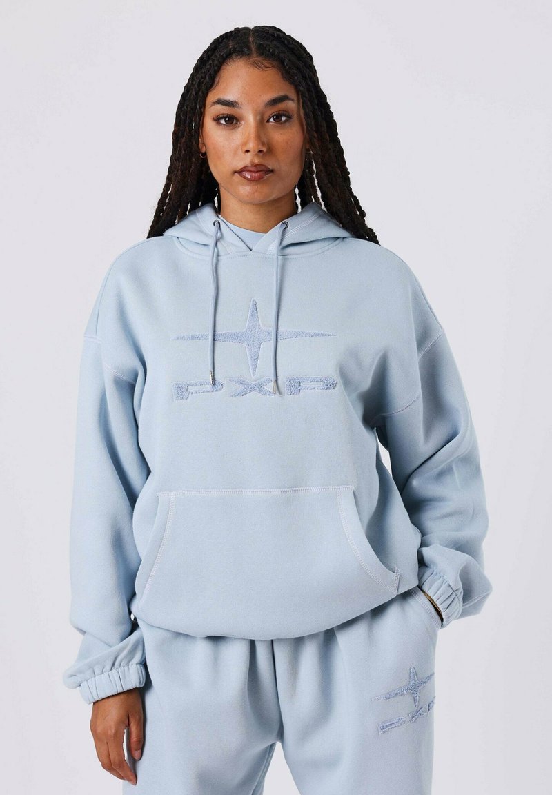 Giovane donna con lunghe trecce, che indossa un hoodie azzurro chiaro e pantaloni della tuta abbinati con il logo "PXP", in piedi davanti a uno sfondo semplice.