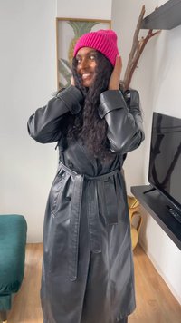 Trench coat in pelle sintetica nera con cintura a fiocco, caratterizzato da ampi risvolti e dettagli con bottoni. Abbinato a un berretto a coste rosa brillante.