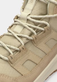 Beige magas szárú sneaker, texturált felsőrésszel, párnázott bokarésszel és lapos fűzőkkel. Suédé díszítések és gumitalp, amelynek jellegzetes mintázata van.