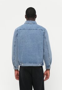 Light blue denim jacket with long sleeves, a collared neckline, and button closures at the cuffs and hem. Simple, textured fabric.