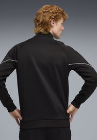 Czarna sportowa kurtka z wysokim kołnierzem, z białymi lamówkami i gładką teksturą. Posiada ściągacze na dole i przy rękawach, zaprojektowana do aktywnego użytkowania.