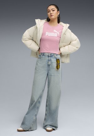 Jachetă tip puffer în crem, tricou roz ajustat cu logo-ul „PUMA”, blugi de dimensiuni mari de culoare albastră deschisă, ochelari atârnând de bucla curelei, încălțăminte maro.