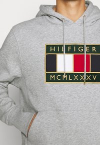 Muž v svetle šedé mikině s kapsou na přední straně a výšivkou loga Tommy Hilfiger, které má černé, bílé a červené pruhy a římské číslice.