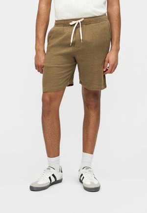 Άνδρας με καφέ υφασμάτινο παντελόνι με κορδόνια, λευκές κάλτσες και λευκά αθλητικά παπούτσια με μαύρες ρίγες, στέκεται μπροστά σε απλό φόντο.