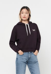 Hoodie violet foncé avec une poche kangourou, des cordons blancs et un logo brodé. Associé à un jean en denim bleu clair.