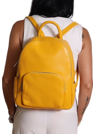 Zaino in pelle gialla con tasca frontale con zip indossato da una persona in top senza maniche bianco e pantaloni, visto da dietro su uno sfondo bianco.