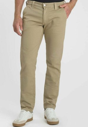 Uomo che indossa pantaloni beige a gamba dritta e sneakers bianche, in piedi con una mano in tasca su uno sfondo chiaro uniforme.