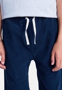 Dziecko ubrane w granatowe sztruksowe spodnie ze sznurkiem w kolorze białym oraz jasnoszarą koszulkę, ręce swobodnie opuszczone wzdłuż ciała, na lewym nadgarstku opaska.