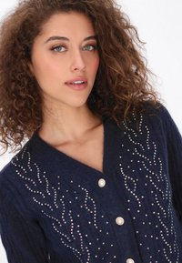 Cárdigan de punto en color azul marino con escote en V, adornado con tachuelas metálicas decorativas y botones de perla, con detalles de costura texturizada y ondulada.