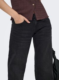 Personne portant un jean noir à jambes larges et un top marron texturé avec des boutons, une main dans la poche et un sac noir visible.