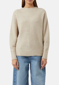 Beżowy sweter w prążki wykonany z miękkiego materiału dzianinowego. Posiada okrągły dekolt i długie rękawy. Połączony z jasnoniebieskimi spodniami o szerokich nogawkach.
