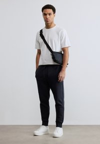 Άνδρας που στέκεται φορώντας λευκό μπλουζάκι, μαύρο παντελόνι τύπου jogger, λευκά αθλητικά παπούτσια και μαύρη τσάντα χιαστί μπροστά σε απλό ανοιχτό φόντο.