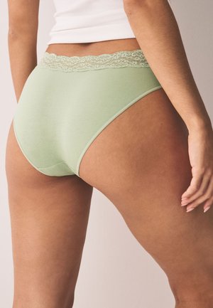 Femme portant un sous-vêtement bikini vert clair avec une ceinture en dentelle et un débardeur blanc, vue de dos et de côté.