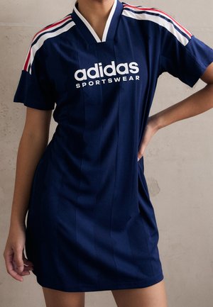 Persoon draagt een marineblauwe Adidas sportjurk met witte en rode strepen op de schouders, poseert met een hand op de heup.