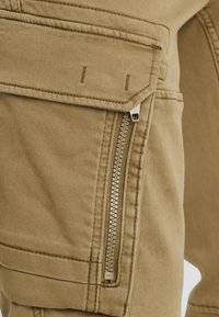 Pantalons cargo khaki avec une poche latérale zippée, des coutures surpiquées, une texture lisse et un gros plan sur le tissu et les détails matériels.