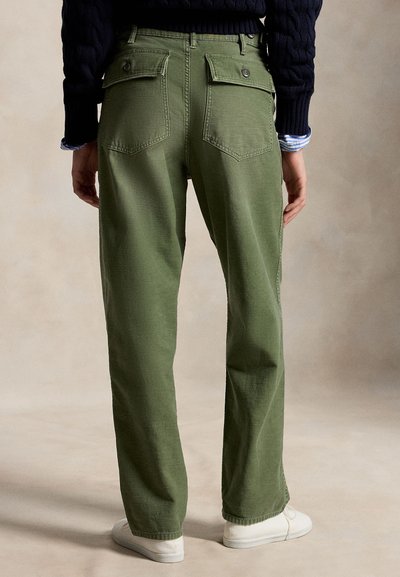 Persoană purtând pantaloni drepți verde măsliniu cu buzunare aplicate cu nasturi în partea din spate, pulover bleumarin și adidași albi, în picioare în fața unui fundal neutru.