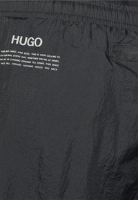Schwarze leichte Shorts mit elastischem Taillenbund und Seitentasche. Auf der Vorderseite farblich bedrucktes "HUGO"-Logo und Textdetails. Textilstruktur.